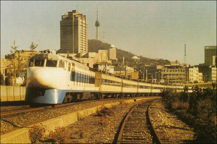 KNR (Korean National Railways) 7100-as sorozatú dízelmozdony (EMD GT26CW) egy 0-s sorozatú shinkansenné „átalakítva”. A járat éppen Szöul állomásról halad ki, háttérben a Seoul Tower és a Namsan Park.