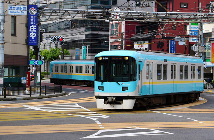Keihan 800-as sorozatú motorvonat Hamaotsu (浜大津) állomás közelében. A társaság Otsu (大津) városában létesített vonalainak érdekessége, hogy villamoshoz hasonló módon integrálódtak a közutakba, melyek rendszerint középen választják ketté az útsávokat.
