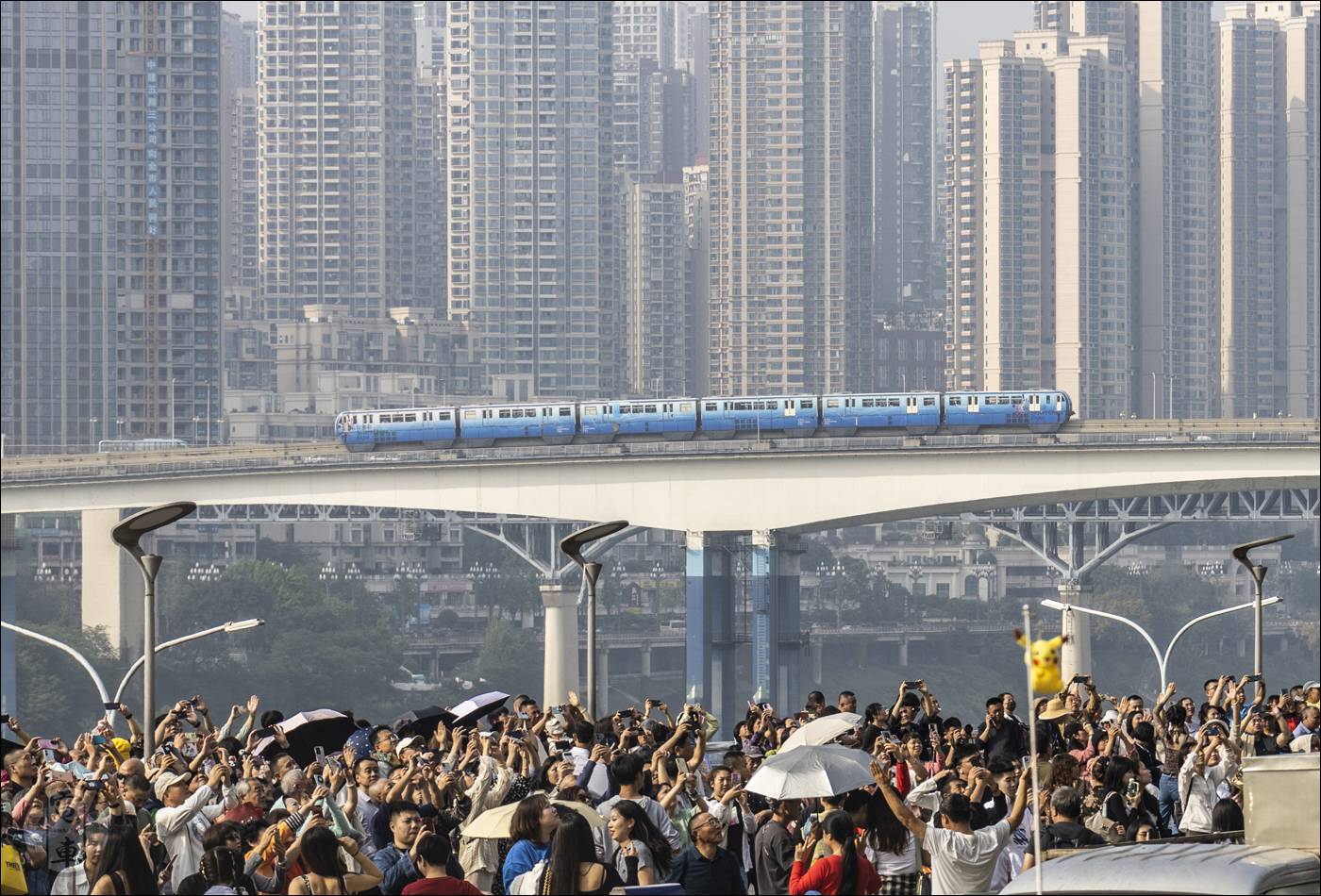 A Chongqing Rail Transit 3-as vonalán közlekedő egyik szerelvény kel át a Jialing-folyó felett, előtérben pedig turisták tömegei örülnek a 2-es vonalon található Liziba megállóba érkező járműnek.