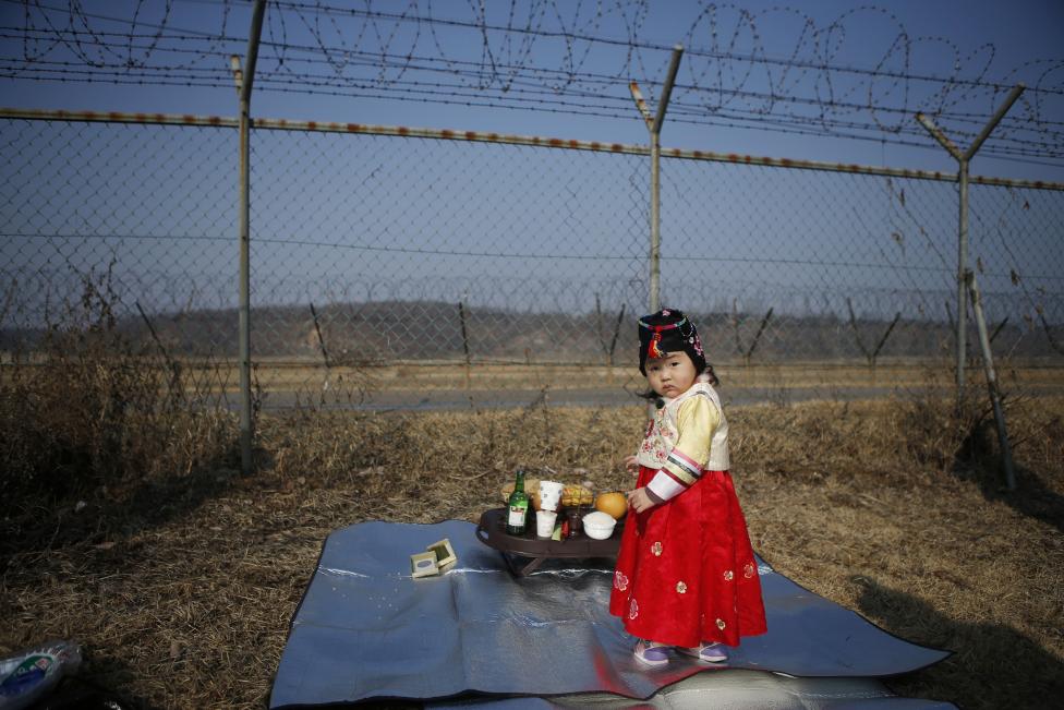 Egy dél-koreai lány áll népi ruházatban a két Korea közti demilitarizált zóna szögesdrót kerítése előtt, miközben szülei előkészítik az észak-korai családtagok tiszteletére rendezett megemlékezést (f.: AFP)