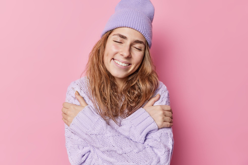 tender-delicate-smiling-young-woman-embraces-herself-with-love-enjoys-comfort-knitted-sweater-keeps-eyes-closed-wears-hat-poses-against-pink-background-has-nice-romantic-memories-feels-soft_wayhomestudio_freepik.jpg