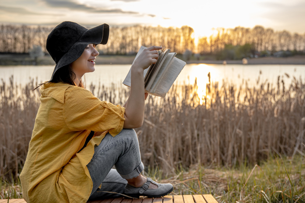 young-woman-hat-with-smile-her-face-is-reading-book-sitting-by-river-sunset.jpg