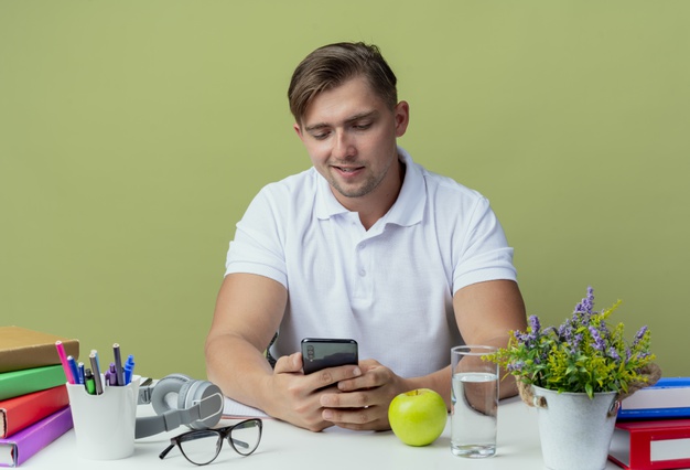pleased-young-handsome-male-student-sitting-desk-with-school-tools-holding-looking-phone-isolated-olive-green_141793-83968.jpeg