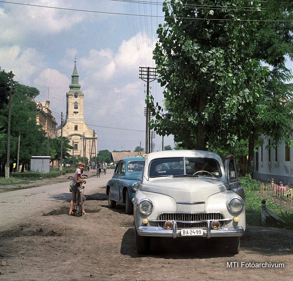 1965. augusztus 14. Kisfiú áll biciklijével parkoló autók mellett - az előtérben egy Warszawa M201, mögötte Moszkvics 403-as taxi - Füzesgyarmat egyik utcáján. Háttérben a református templom. MTI Fotó: Kunkovács László