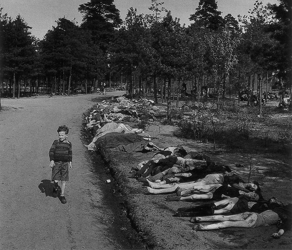 1945. Német fiú a felszabadított bergen-belseni koncentrációs táborban a kivégzettek sorfala előtt, rezzenéstelenm arccal sétál. A háttérben a fák között is hullahegyek..jpg