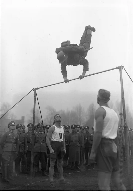 1934_a_german_soldier_completing_a_giant_wave_on_a_horizontal_bar_in_full_marching_kit.png