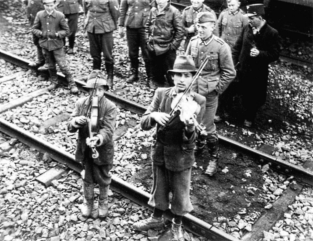 1943_ket_cigany_fiu_hegeduszoval_fogadja_a_francia_beaune-la-rolande_atmeneti_taborba_erkezo_zsidokat_a_taborbol_auschwitzba_mentek_a_transzportok_a_hatterben_nemet_katon.jpg