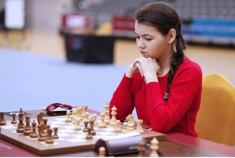 aleksandra-goryachkina-with-braided-pony-tail-in-red-sweater-sitting-at-chessboard-behind-whites-pieces.jpg